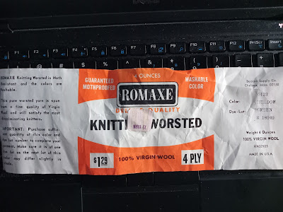 A detached yarn label lays across a computer keyboard. The label is white with an orange block design in the center where the ROMAXE logo sits above the name of the yarn. It is clearly very old.