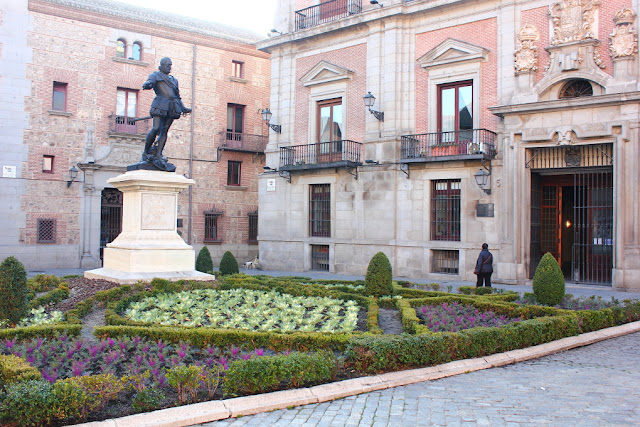 Na placyku Plaza de la Villa w Madrycie. W tle ceglane, renesansowe budynki o kamiennych boniowaniach naroży, dekoracyjnych portalach i obramowaniach okien. Przed nimi ustawiona na cokole brązowa rzeżba Alvaro de Bazan. Przy rzeźbie kwiatowa rabatka. 