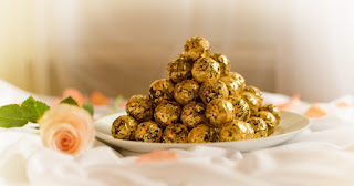 Descrição: foto em fundo desfocado. Ao centro, sobre um tampo forrado por tecido branco e fofo, um prato de louça branca, dentro, uma pirâmide de bombons dourados. Detalhe: o suíço DeLafée é preparado com grãos finos de cacau, açúcar, óleo de coco, manteiga de cacau, leite e baunilha. Até aí tudo bem. O diferencial está na cobertura: flocos comestíveis de ouro 24 quilates, aplicados à mão sobre cada bombom. À esquerda do prato, uma rosa salmão e ao redor, algumas pétalas decoram o momento.  Com esta imagem desejamos um doce e Feliz Dia dos Pais!