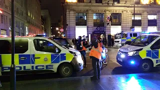 Night street in London. Three yellow and blue police vans are parked. Between the two vans in the front there are some people standing including reporters with microphones and cameramen