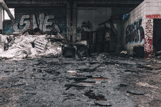 Desolate scene inside a dilapidated structure with graffiti-covered walls, charred debris, and a black liquid on the floor.