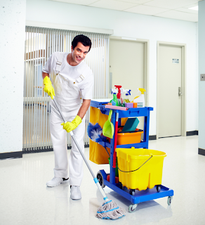 Photo of a dark-haired male janitor in a white outfit mopping the floor of a building with his janitorial supplies nearby.