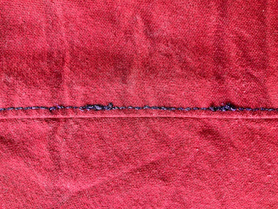 a close up of dark red fabric with a line of decorative topstitching running along a seam. The topstitching is done in a sparkly, dark purple thread using a ~4mm stitch. There are several places where the thread has broken and had to be re-sewn. End ID.