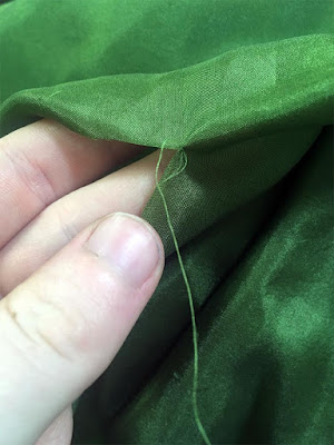 A close-up of a neat slit in thin green silk, held open by a glowing white hand, with a very fine thread emerging from the end of the slit.