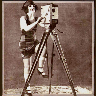 Sepia-toned black and white photo of silent film actress Charlotte Rich in a bathing suit posed as if she's cranking a movie camera. From page 1 of the February 11, 1922 National Police Gazette.