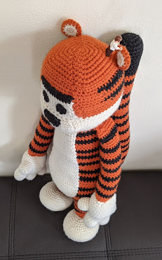 A crochet Hobbes (tiger) plushie standing up, with photo from above. A lego man is standing next to the ear, which is about the same size. This is to show the scale of the toy as it's very large.