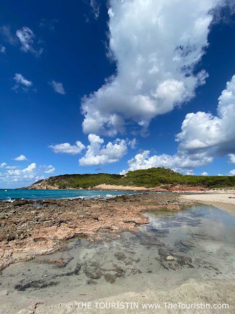 Rockpools on the shore of a white sandy beach with turquoise water, and lined by sandstone rocks and lush greenery under a blue sky with fluffy white clouds.