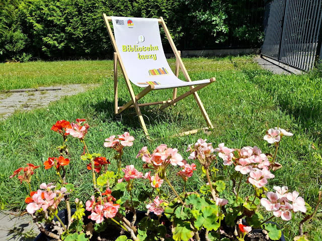 Leżak na tle zieleni. Przed leżakiem stoi doniczka z kwiatami.