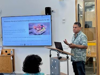 A man of Australian Aboriginal decent is delivering a presentation in front of a large screen. He is standing at a lectern, moving his arms as he discusses a point on the slide. The screen shows a slide illustrating a Mbabaram knowledge-based drug that could be used to treat IBD.