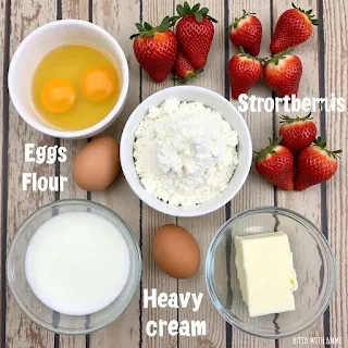 "Ingredients for Strawberry Shortcake Cake Roll, including eggs, flour, sugar, fresh strawberries, and heavy cream, arranged on a wooden table.