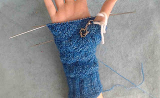 someone wearing a glove in progress, knit in fingering-weight blue yarn. A stitch marker with a silver dolphin charm is clipped into the work.