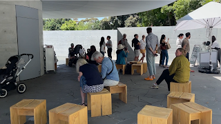 Short wooden stools with people sitting on them on a grey concrete floor with grey concrete walls and an open ceiling.