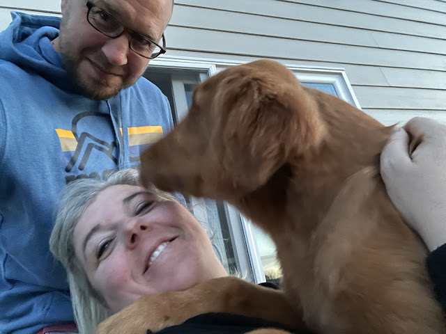 A selfie of me and dad in the backyard on the patio with Miles in my lap as I am sitting down in a chair (dad is standing behind me). Miles is licking my face.