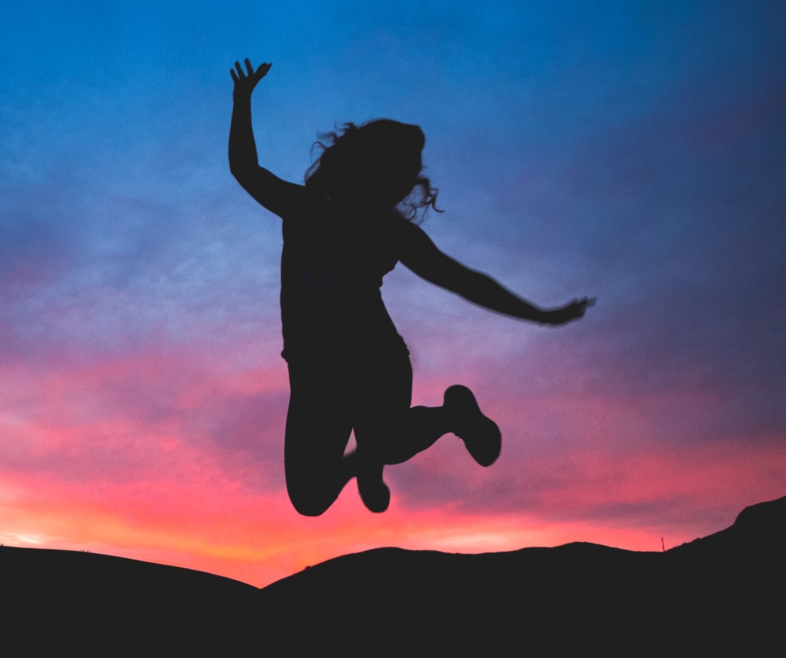 Photo of a silhouette of a person jumping in the air, arms outstretched, legs bent up behind them, against the backdrop of hills in silhouette and a orange, pink, purple and blue sunset