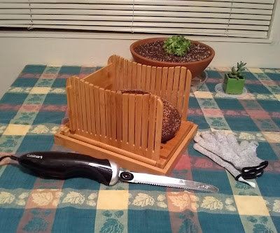 On a table-top that's covered with an oak-leaf patterned tablecloth in colors of green, brown and gold: an electric bread knife, loaf of bread inside a cutting board that has slatted sides, and a pair of knit, grayish-silver gloves. Behind them are a couple of succulents in pots.