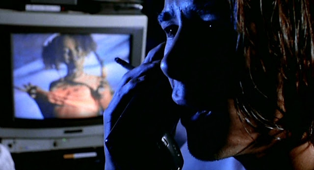 A film still from Nowhere. A man is on the phone. In the background, there is a woman on TV.