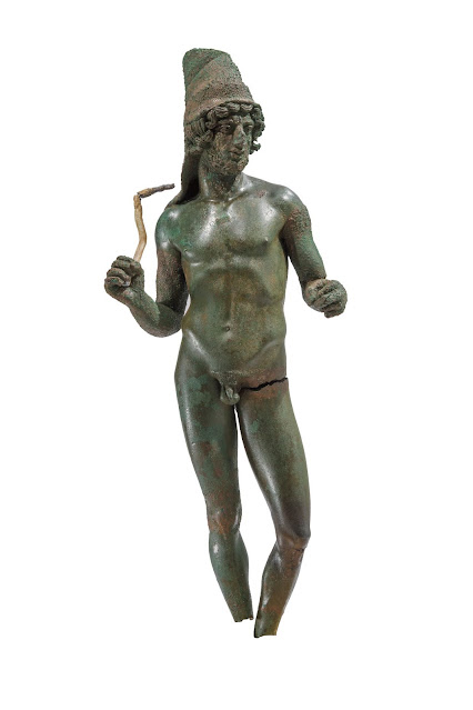 Χάλκινο αγαλμάτιο γυμνού νέου που κρατά όρθιο μολύβδινο ραβδί στο δεξί του χέρι. Φρυγικός σκούφος καλύπτει τον αυχένα και την ωμοπλάτη. Ρωμαϊκή Περίοδος. [Credit: Σ. Μαυρομμάτης]