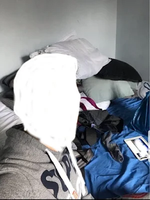 Random guy from a gay dating app doing a selfie with his very messy bed with lots of clothes and other junk in the background.