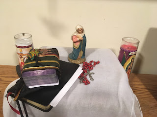 an image of my nightstand: a small statue of the Virgin Mary, a red rosary, a prayerbook, and a set of tarot cards, all flanked by two votive candles.