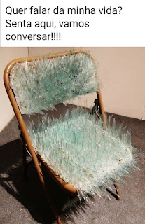 Foto de uma cadeira dobrável aberta, posicionada no canto de um ambiente acarpetado. Cacos grandes de vidros pontiagudos revestem o assento e o encosto da cadeira. No topo lê-se: Quer falar da minha vida ? Senta aqui, vamos conversar!!!!