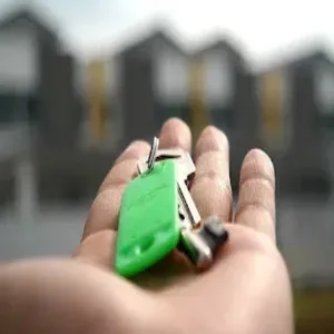 Foto imagem com fundo desfocado de uma casa e  em primeiro planno, uma mão estendida com um molho de chaves sobre esta, representando texto sobre seguros na locação de imóveis.