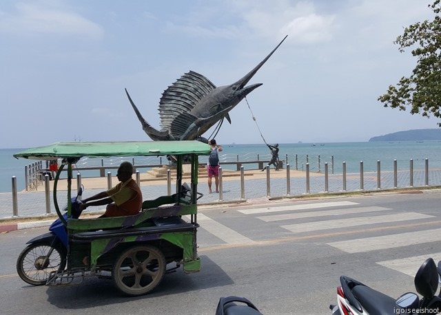 A tuk tuk passing by the swordfish statue along Ao Nang Beach.