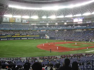 Baseball field in an indoor stadium called the Kyocera Dome in Osaka Japan. The baseball diamond is brown and to the right. The outfield and grass is on the left side of the image. There are stadium seats surrounding the baseball field and bright lights with Japanese advertisements under them.