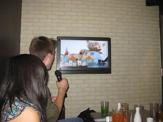 Man and woman on the left side with their back to the camera. Man is holding a microphone. They are looking at a small television on a white brick wall. In front of them is a small table covered in pint glasses and sake containers