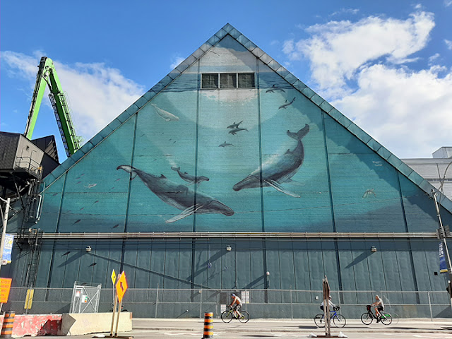 Mural on the side of a peaked industrial building. The mural features whales