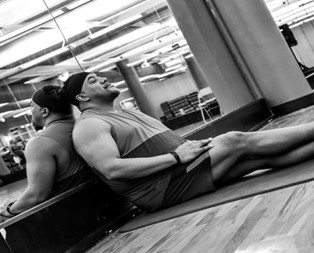 A black and white photo of a man in athletic gear resting against the mirror of a workout studio. This is a portrait of fitness instructor Russell Lee.