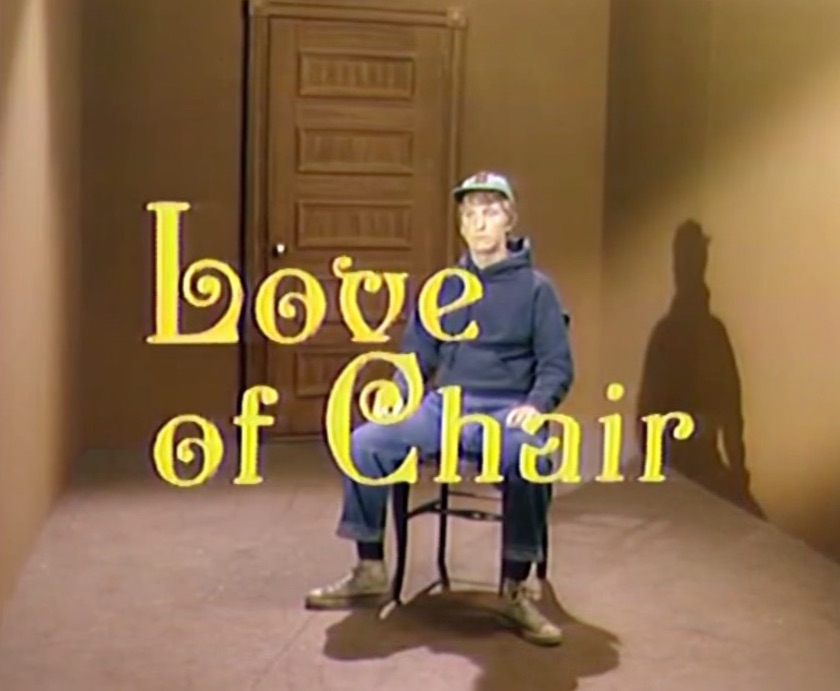 The words 'Love of Chair' over scene of young man sitting on a chair in a small, empty room