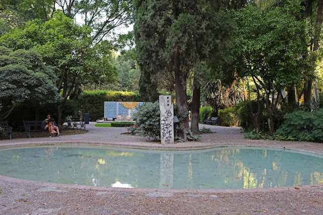 Pequeño lago artificial en la plaza de un parque con frondosa vegetación.