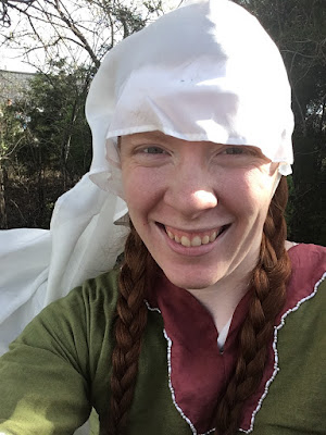 A selfie of a young white woman with long red pigtail braids, in a fluttering white veil and green dress with pink trim, outlined in pearls. Sunlight falls from her left, which is where the wind is blowing from, too.