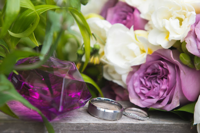 A pair of silver weddings rings surround by flowers and a purple crystal.