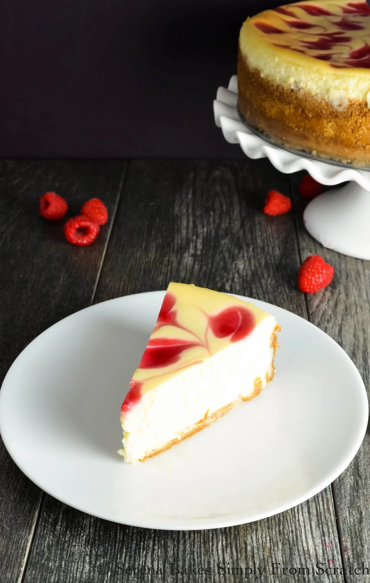 A side shot of a Tall Creamy Cheesecake with Raspberry Swirl slice on a white plate with a whole Cheesecake in the background on a white cake stand.