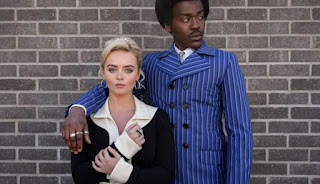 The Doctor (Ncuti Gatwa) and Ruby Sunday (Millie Gibson) in Swinging London apparel (the Doctor's a blue pin-stripe suit), against a brick wall. The Doctor looks off to the left, Ruby looks at the camera.
