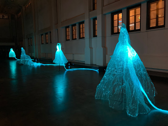 Ghostly dresses made of a fine glowing mesh form a line in a large room. Each has a glowing umbilical cord like structure attached to it, running away along the floor.