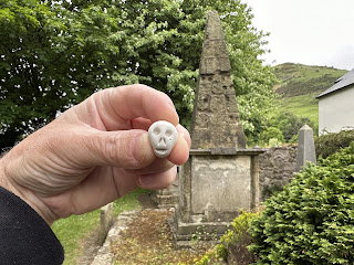 A photo of a hand holding up a small ceramic skull (Skulferatu 102).  In the background is the rather shabby memorial stone.  Photo by Kevin Nosferatu for the Skulferatu Project.