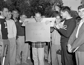 A photograph of a crowd of men. In the middle, a young man is stuck in a stock and holding a sign reading "I wouldn't wear my beanie."