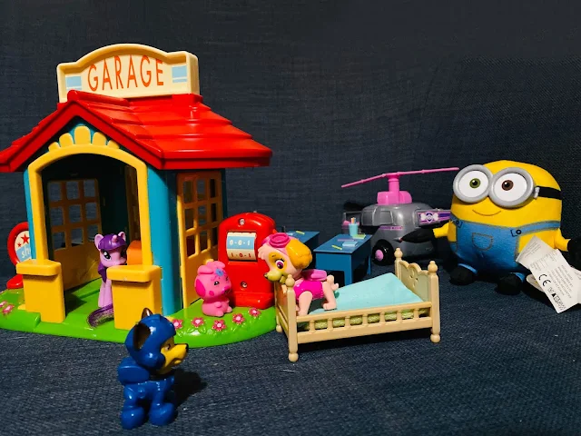 A multicoloured toy world including a Happyland Garage with a my little pony peering out, skye from Paw Patrol standing on a bed and talking to Chase, some Peppa Pig desks in the background next to Skye's helicopter and a soft minion toy