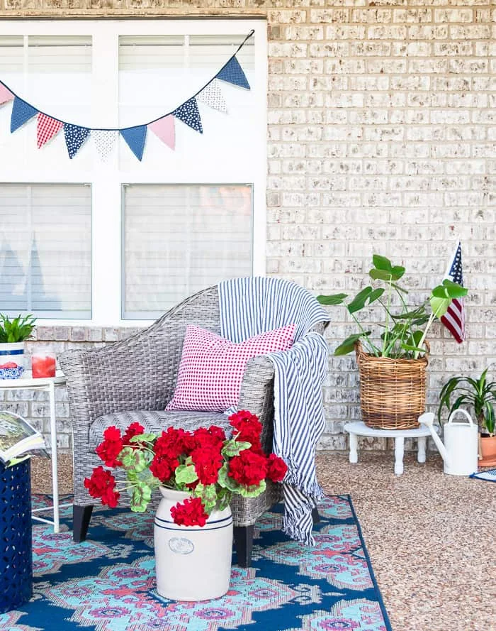 faux red geraniums in vintage crock, wicker chair, blue striped throw, red gingham pillow, greenery