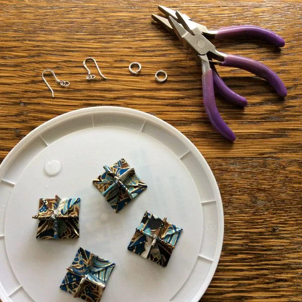 folded paper earring components glued and drying on plastic lid alongside jewelry pliers and silver earring findings