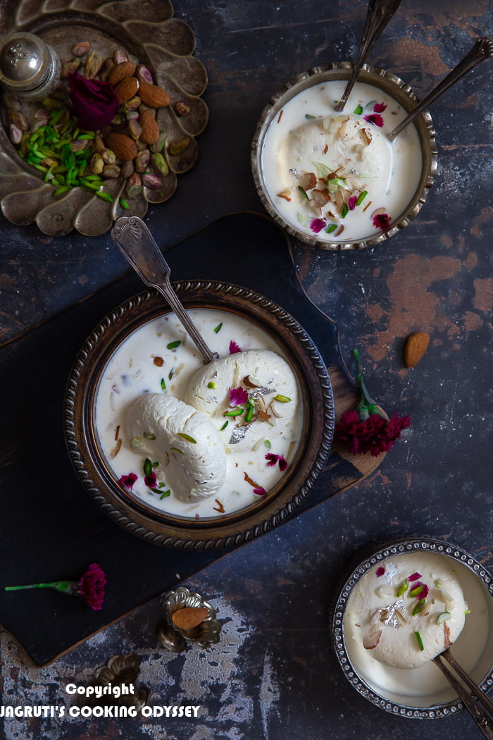 White ras malai served in a metal bowl with a spoon on black chopping board next to pistachio and almond bowl.