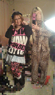 Mirror selfie of Ray and Olivia, Olivia is wearing a black hat, a black long sleeve with monochromatic and pink graphic text across the front, acid washed denim skirt, black and pink star print leggings, and black platform ankle boots.