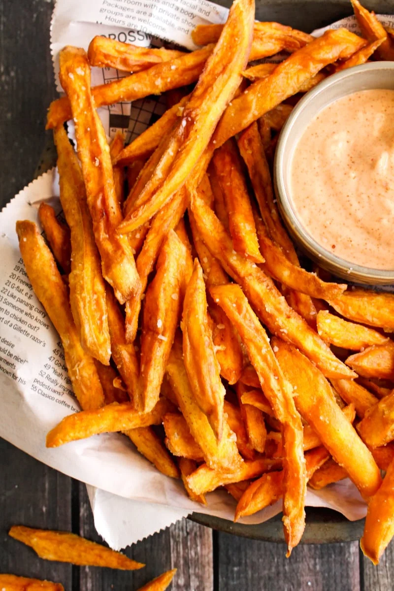 Closeup top view of sweet potato fries on a pie plate lined with newspaper on a wood background.