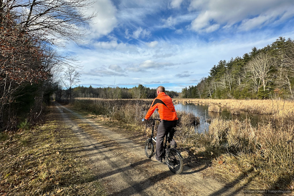 Cyclist wearing an orange jacket on a dirt track, riding past an expanse of water to the right with reeds and trees.
