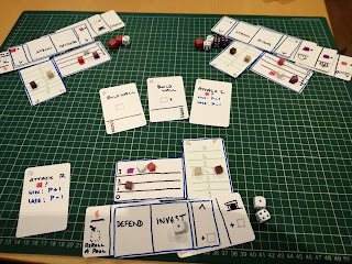 Various hand-written cards arranged in groups, many cards overlapping, on a green mat with a square grid on it. There are also dice and wooden cubes on or near the cards.