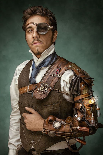 Man wearing Steampunk clothing. He wears a large glass monocle. Clothing: waistcoat/vest, high collar shirt, tie, and steam powered bracer/mechanical arm. Men's steampunk fashion.