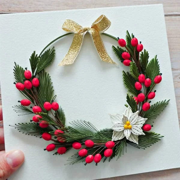 hand holding quilled wreath decorated with red berries, gold bow, and white poinsettia