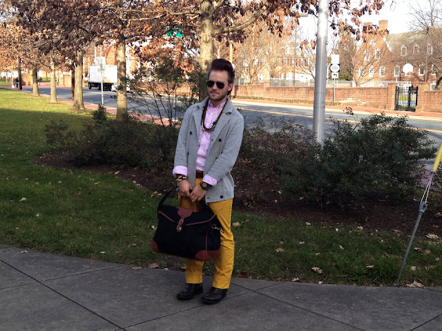 Adam Damien-Lafayette Worton, is a sophomore Apparel Design Major at the University of Delaware. He is wearing a pink button down shirt under a gray notch lapel wool blazer and a pair of light rust chinos. He accessorizes with a wooden necklace and bracelet, a watch and a black and brown detailing bag and black shoes. 